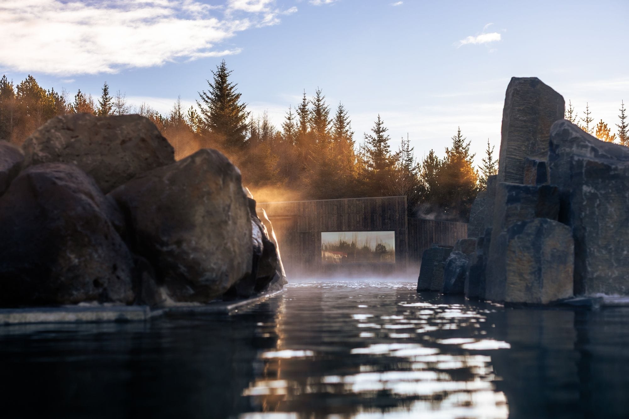 Steam rising from the lagoon between large boulders and basalt columns, with a timber building and spruce trees in low sunlight
