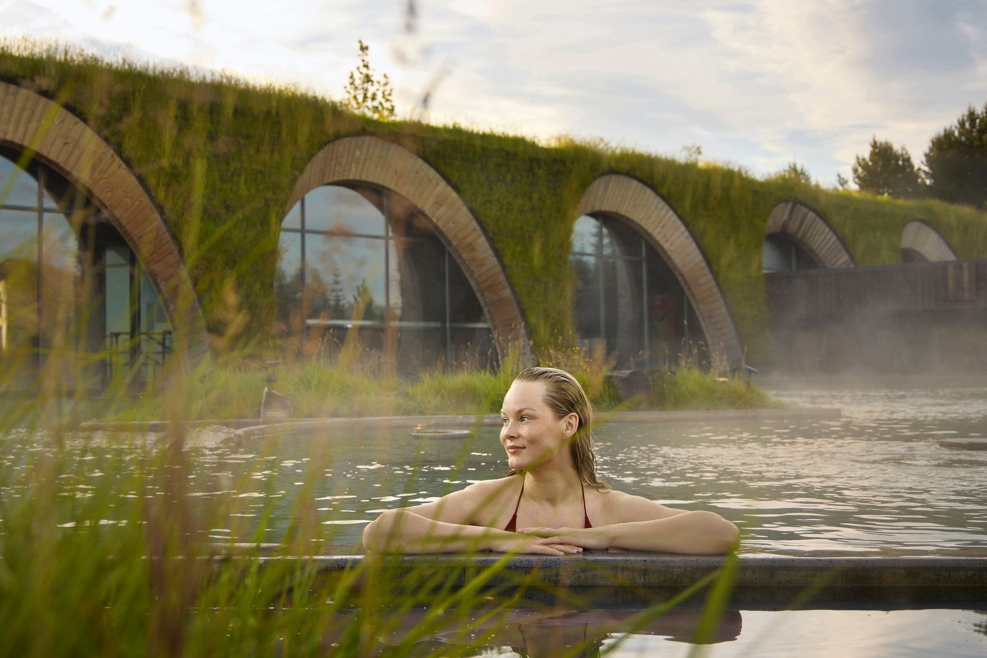 A woman resting her arms on the edge of the lagoon, with the arched, grass-covered building behind her