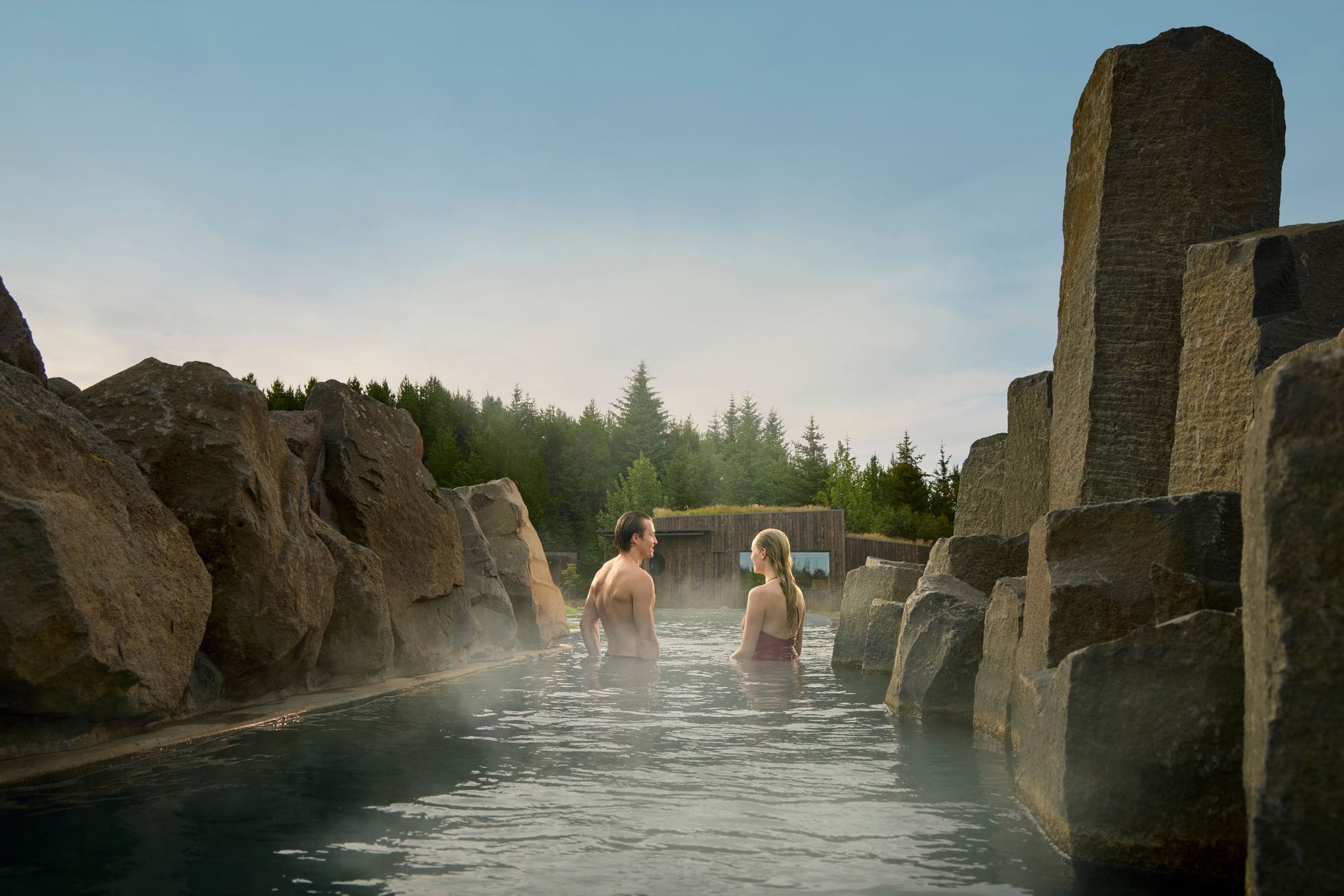 A couple standing in the warm water of a narrow channel between rocks and basalt columns, surrounded by spruce trees