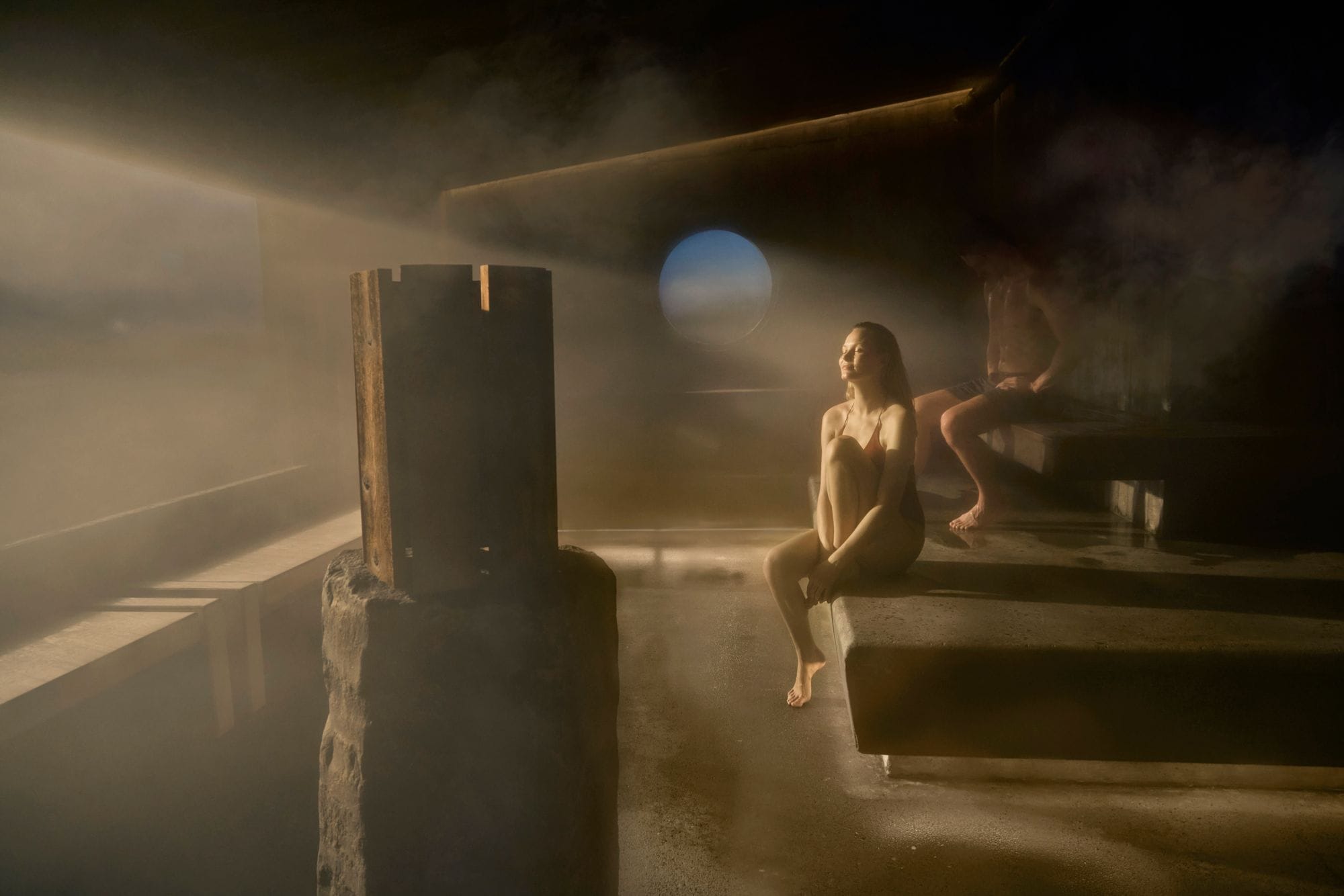 A woman sitting with her eyes closed in the steam room as a beam of light falls through a round window